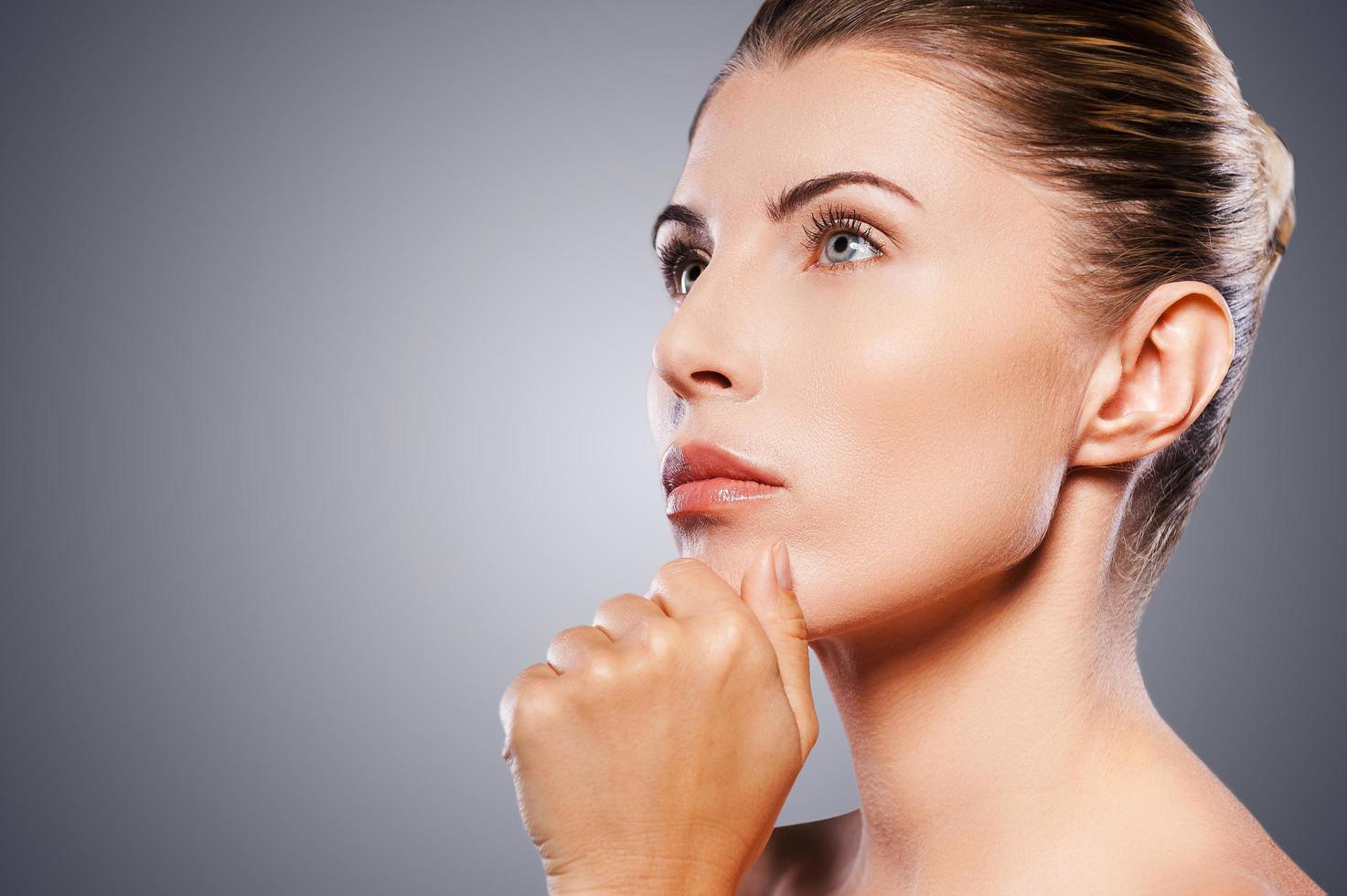 Thoughtful mature beauty. Side view of thoughtful mature woman holding hand on chin and looking away while standing against grey background photo