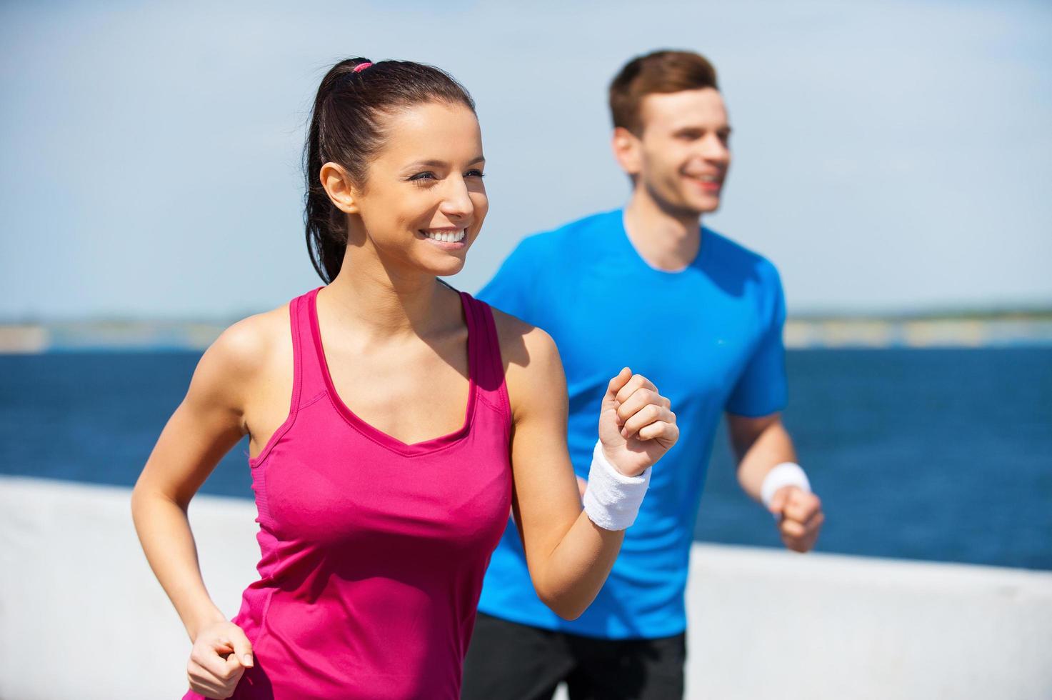 el deporte es nuestra vida. alegre joven y hombre con ropa deportiva corriendo a lo largo de la orilla del río y sonriendo foto