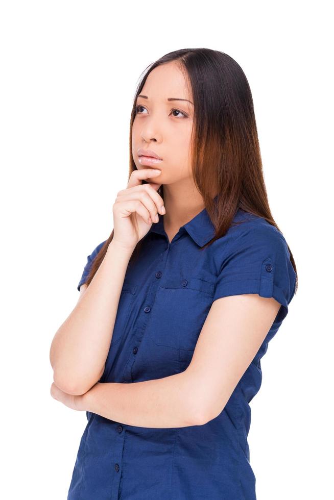 Lost in thoughts. Thoughtful young Asian woman holding hand on chin and looking away while standing isolated on white photo