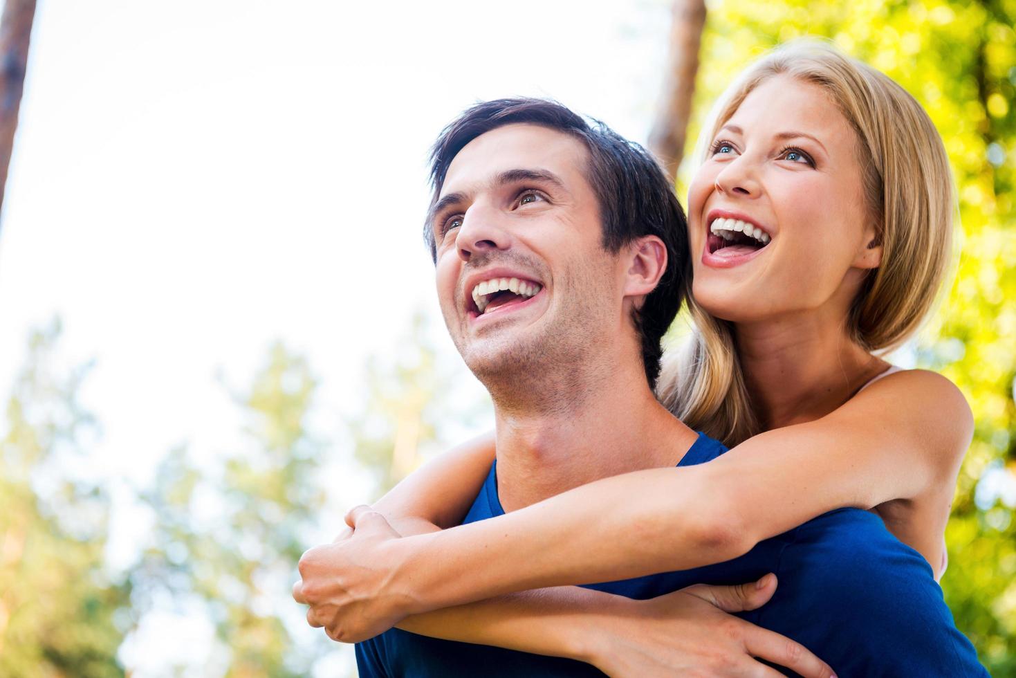 Happy to be together. Low angle view of happy young loving couple standing outdoors together while woman hugging her boyfriend from back and smiling photo