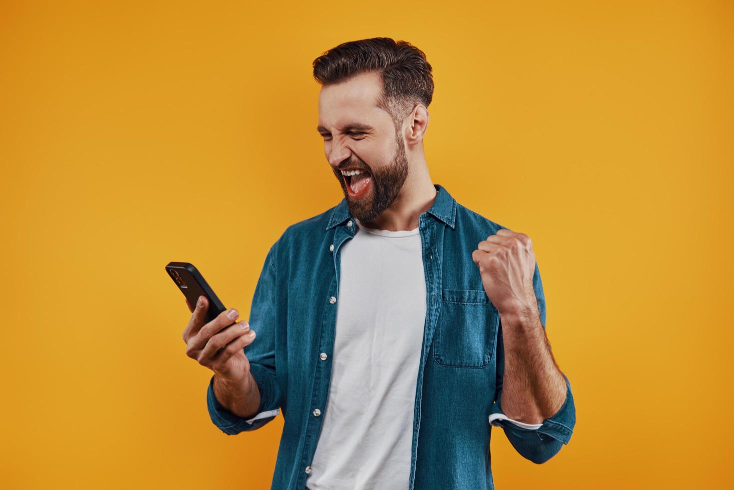 Charming young man in casual clothing holding smart phone and cheering while standing against yellow background photo