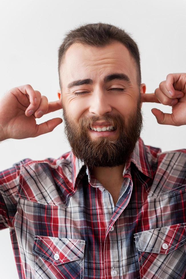 no escuchar el mal. apuesto joven barbudo haciendo una cara y cubriendo las orejas con los dedos mientras está aislado en un fondo gris foto