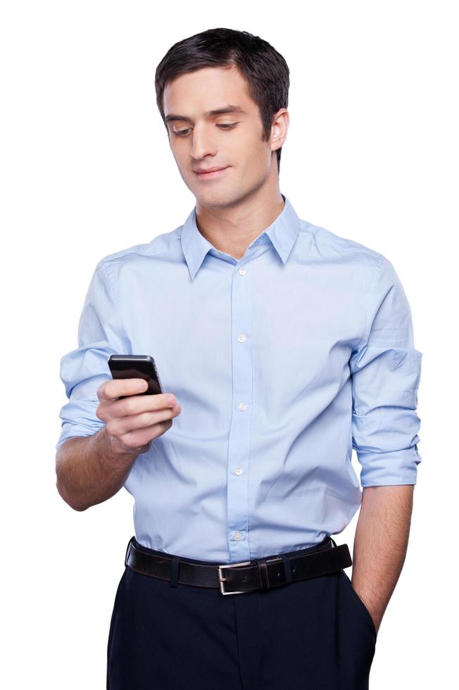 Always in touch. Handsome young man in formalwear holding mobile phone and smiling while standing isolated on white photo
