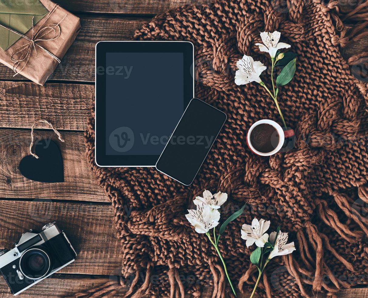 Modern technologies. High angle shot of knitted scarf, flowers, cup of coffee, gift box, photo camera, smart phone and digital table on wooden desk