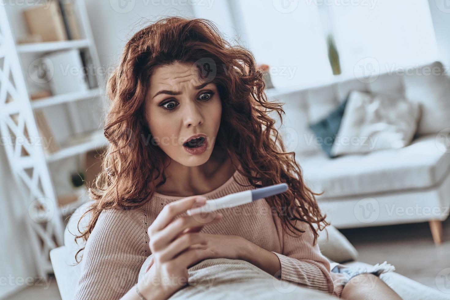 No way Terrified young women looking at pregnancy test and keeping mouth open while sitting on the sofa at home photo