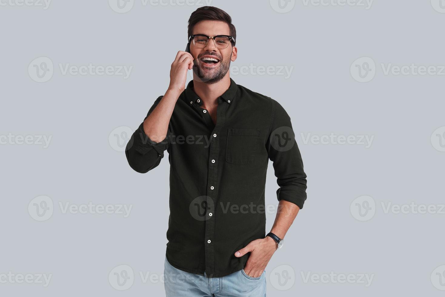 Good talk with client. Good looking young man in eyeglasses talking on smart phone and smiling while standing against grey background photo
