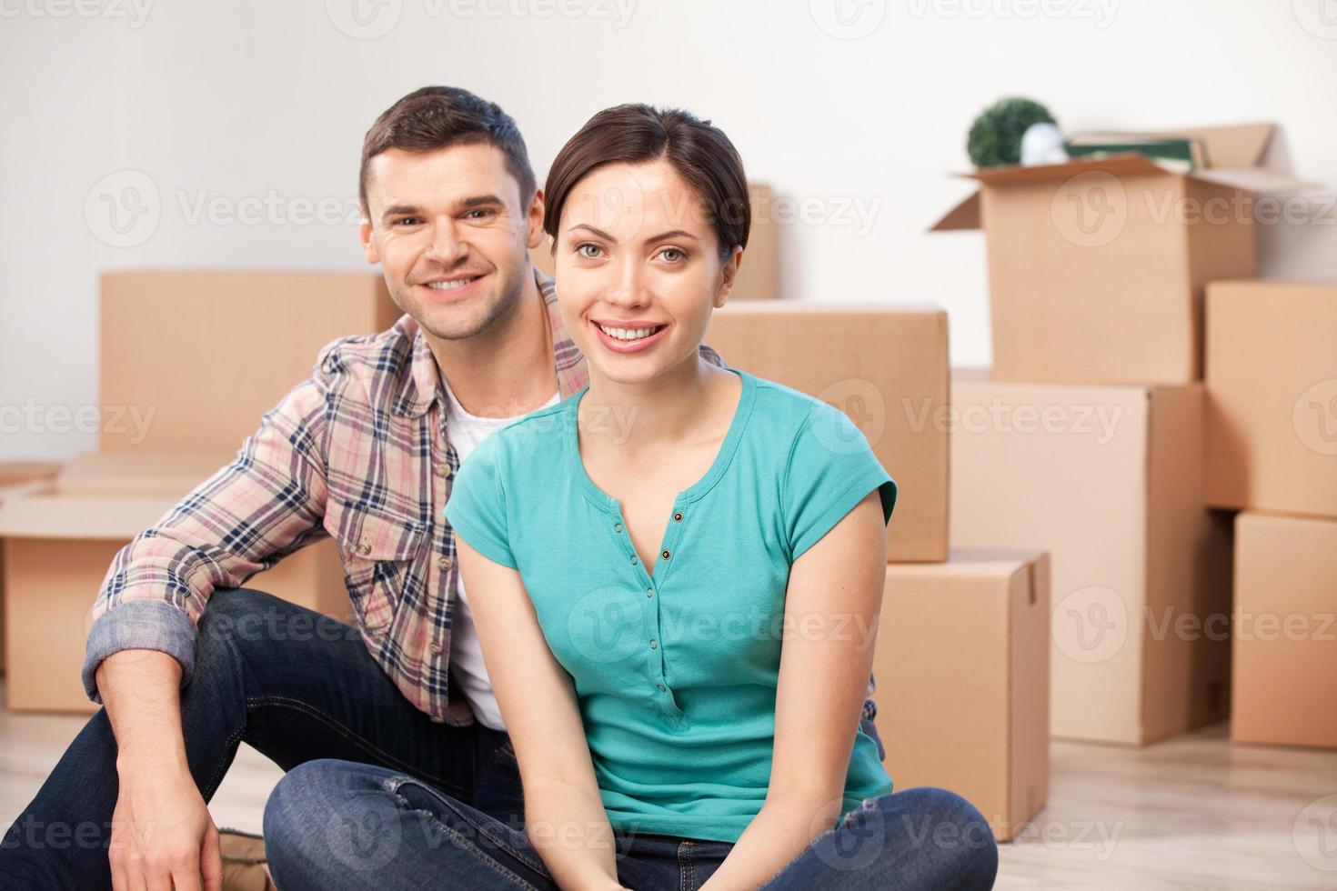 Together in a new apartment. Cheerful young couple sitting close to each other and smiling while cardboard boxes laying on background photo