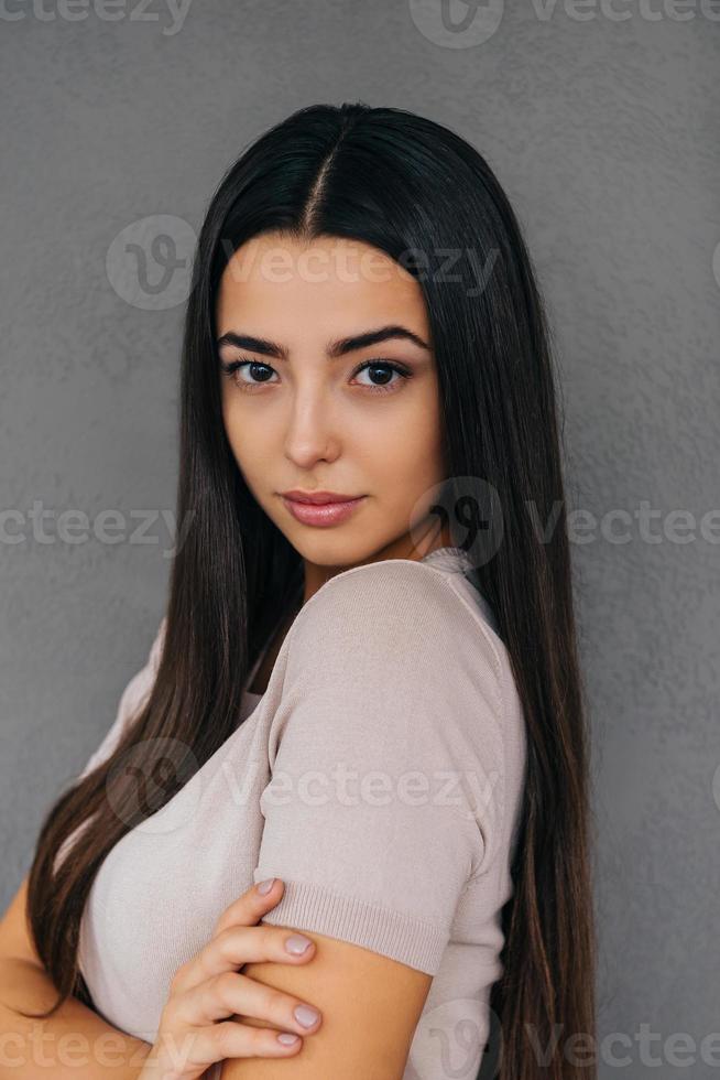 confiado en su belleza. hermosa mujer joven con los brazos cruzados y mirando a la cámara mientras está de pie contra el fondo gris foto