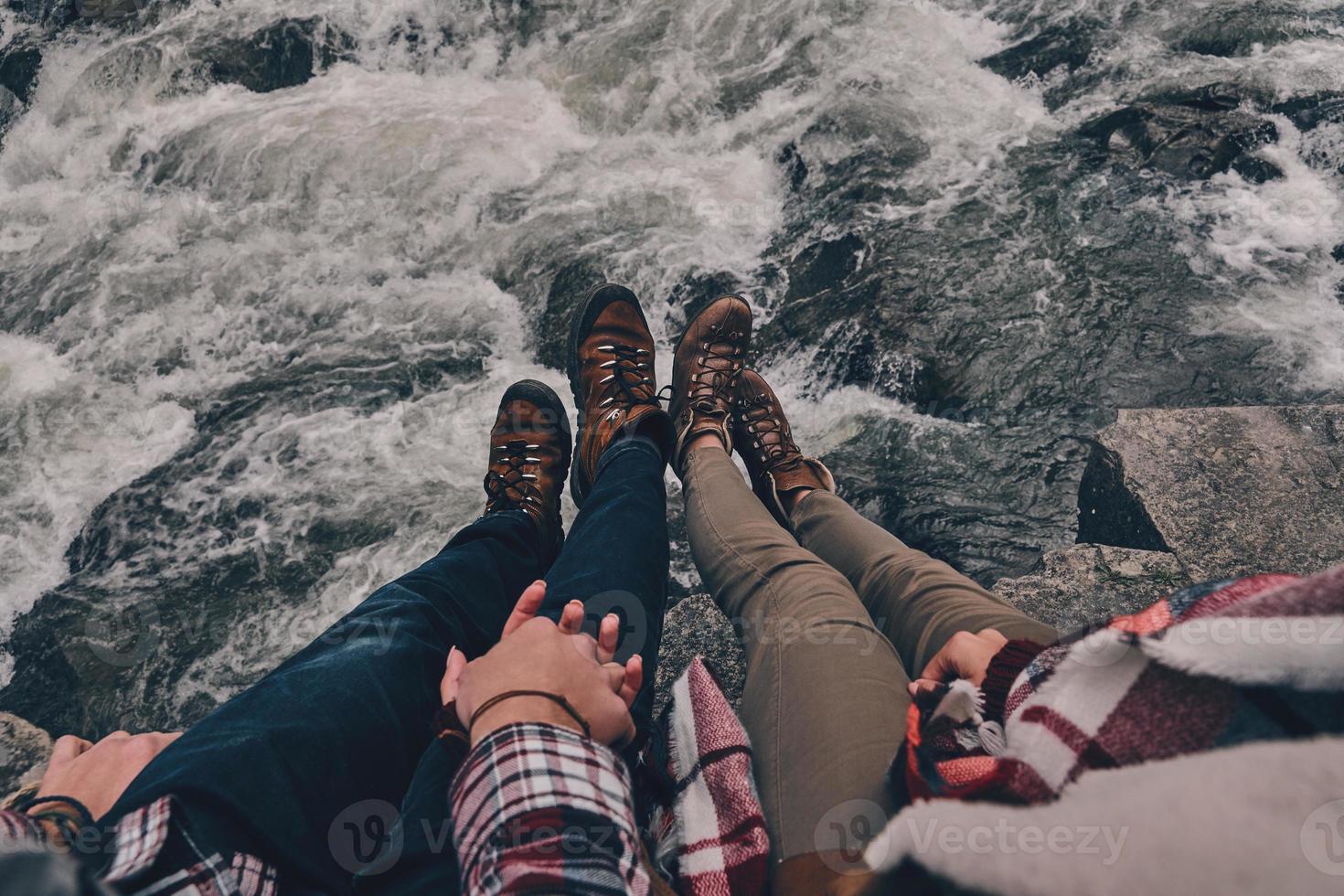 Always together. Close up top view of young couple holding hands while sitting on the rocks with the river below photo