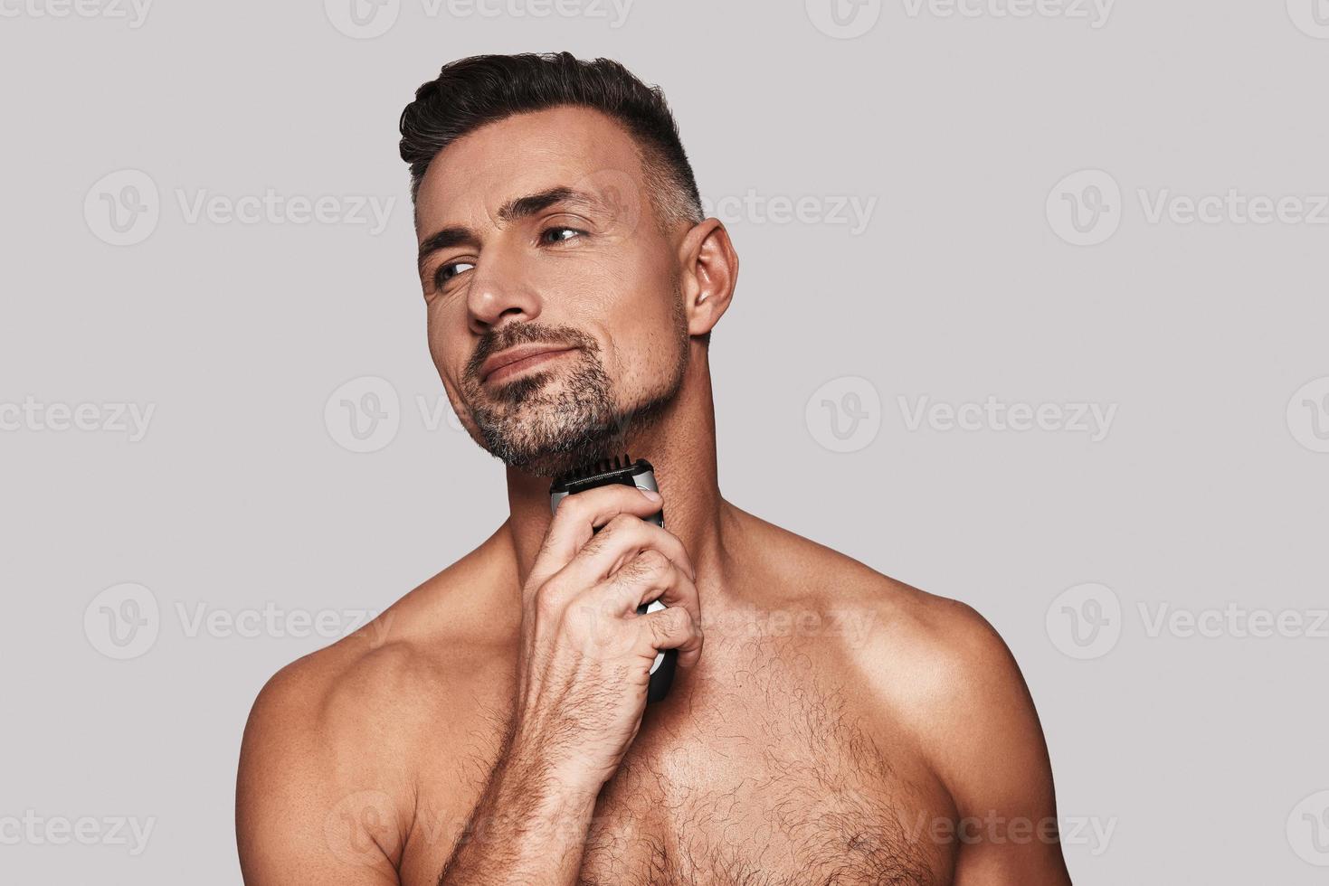 Man shaving. Charming young man shaving and smiling while standing against grey background photo