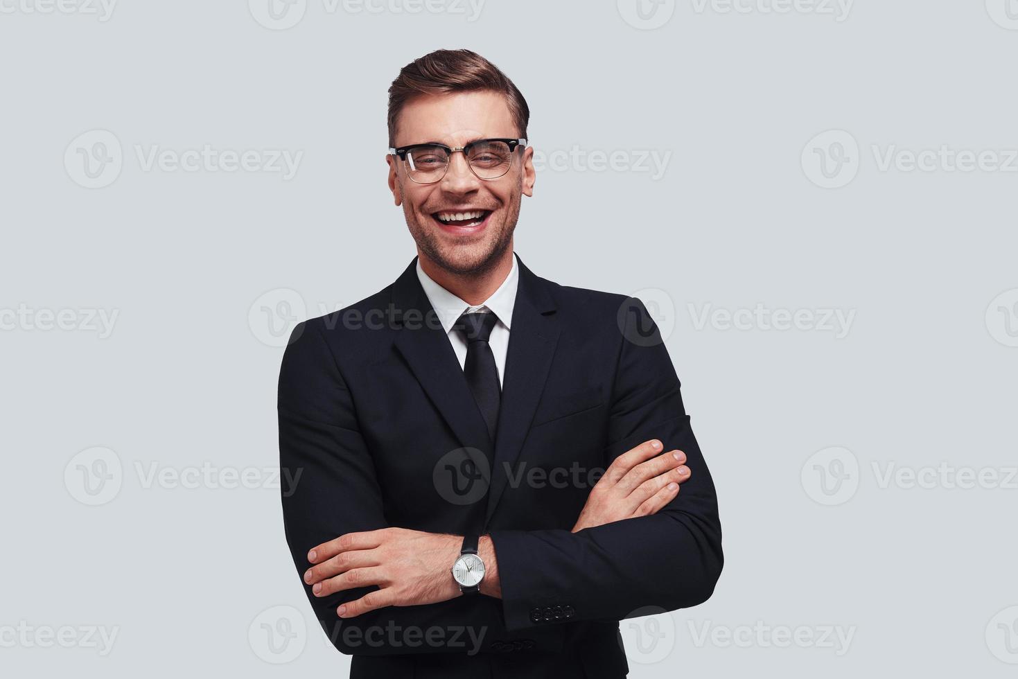 Used to look perfect. Handsome young man in full suit keeping arms crossed and looking at camera with smile while standing against grey background photo