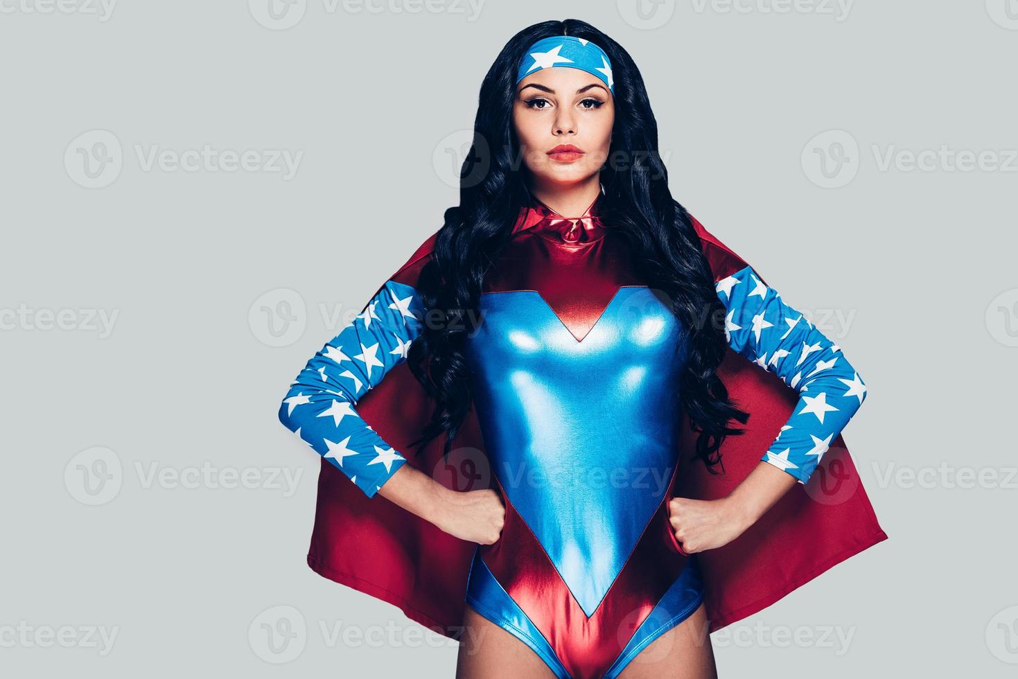 She can fight crime anywhere. Beautiful young woman in superhero costume looking at camera and keeping hands on hips while standing against grey background photo