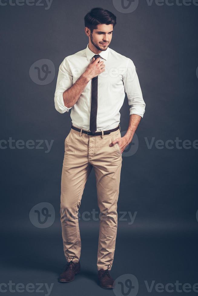 Thoughtful handsome. Full length of confident young handsome man adjusting his tie and looking away while standing against grey background photo