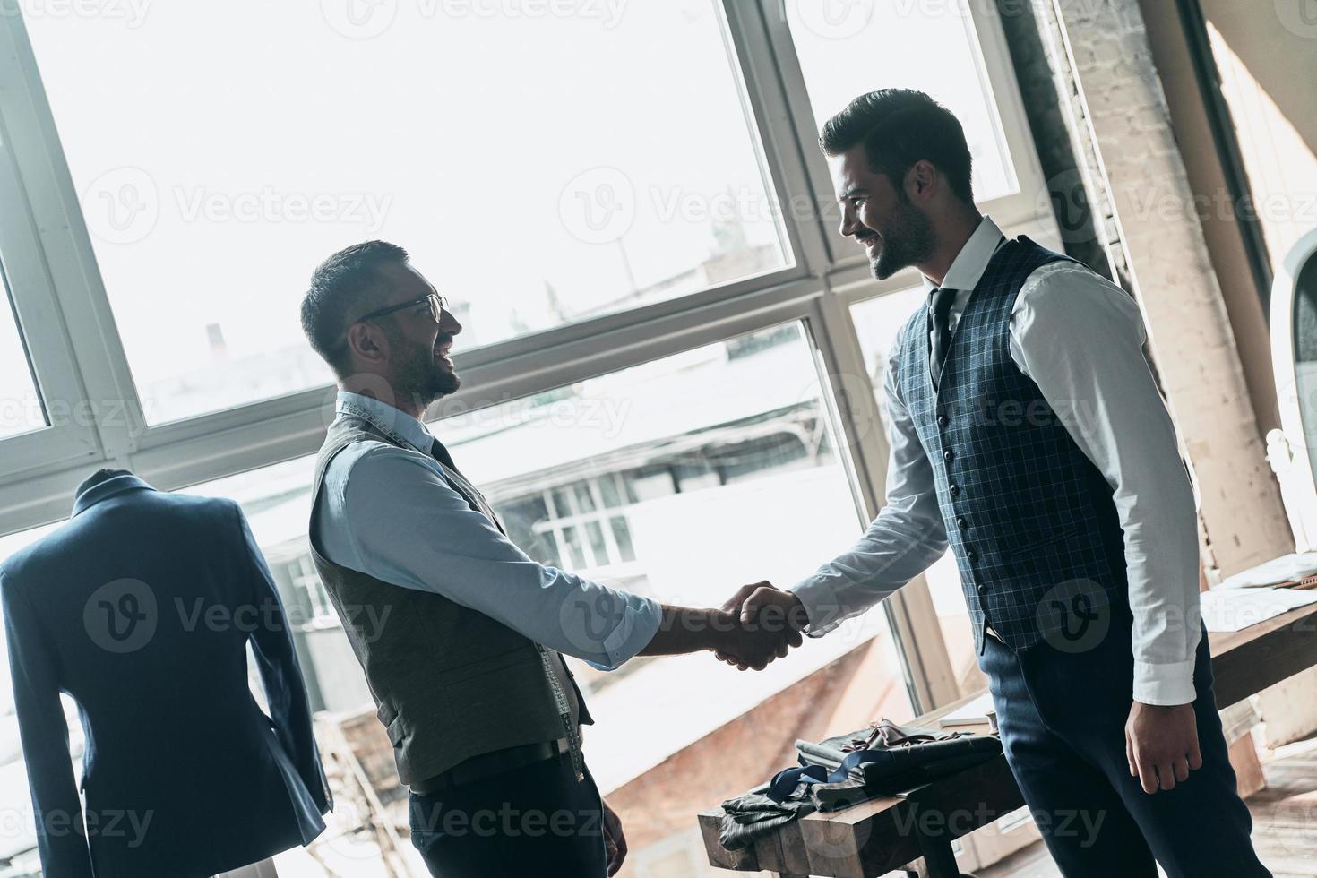 Two young fashionable men shaking hands and smiling while standing in workshop photo