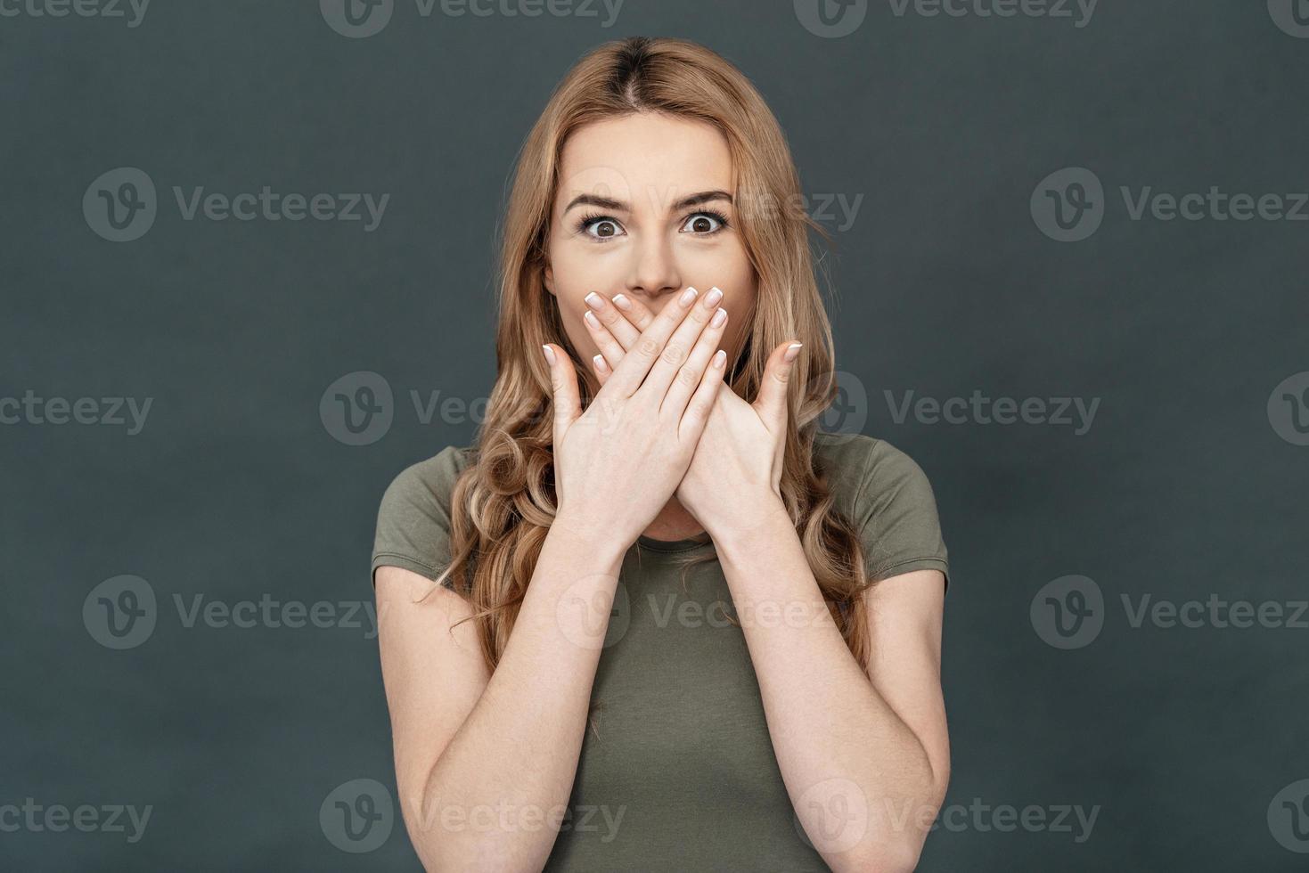 Trying to stop gossiping. Surprised young woman in casual wear covering mouth with hands and looking at camera while standing against grey background photo