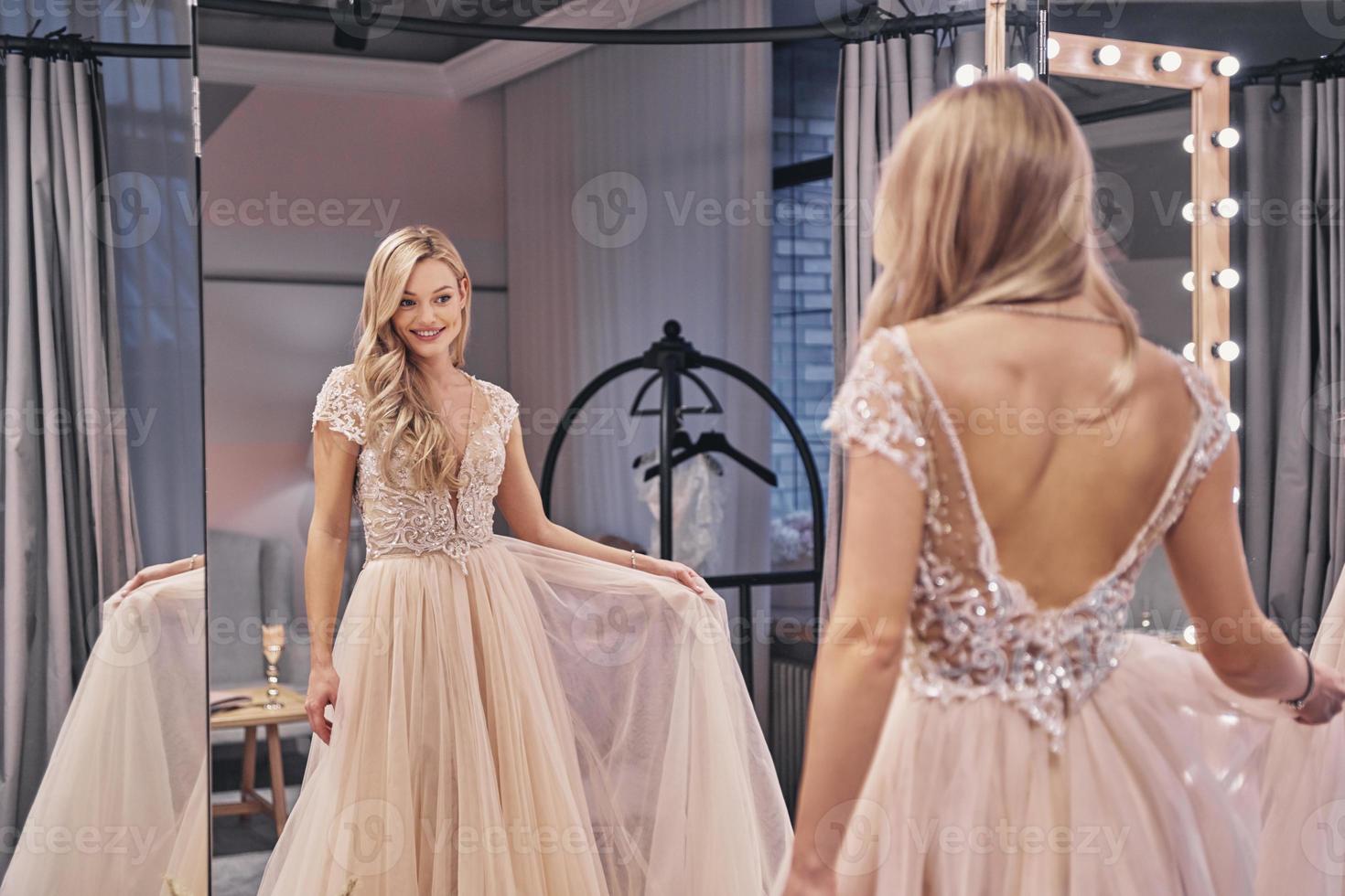 What a lovely dress.  Beautiful young woman wearing wedding dress and smiling while standing in front of the mirror in bridal shop photo