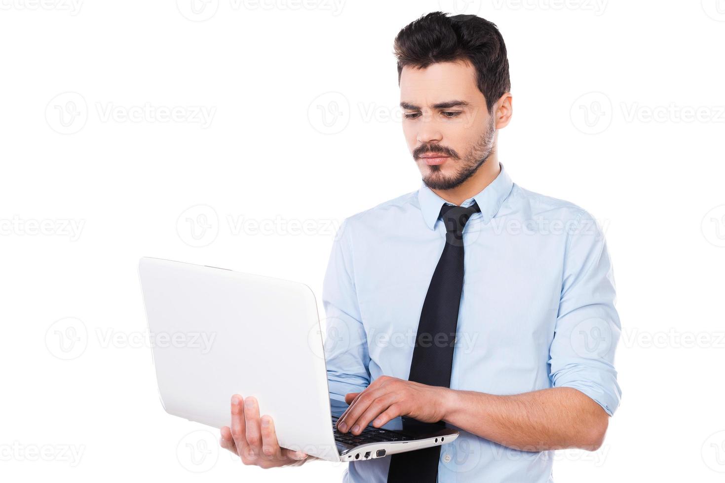 Confident business expert. Handsome young man in shirt and tie working on laptop while standing against white background photo