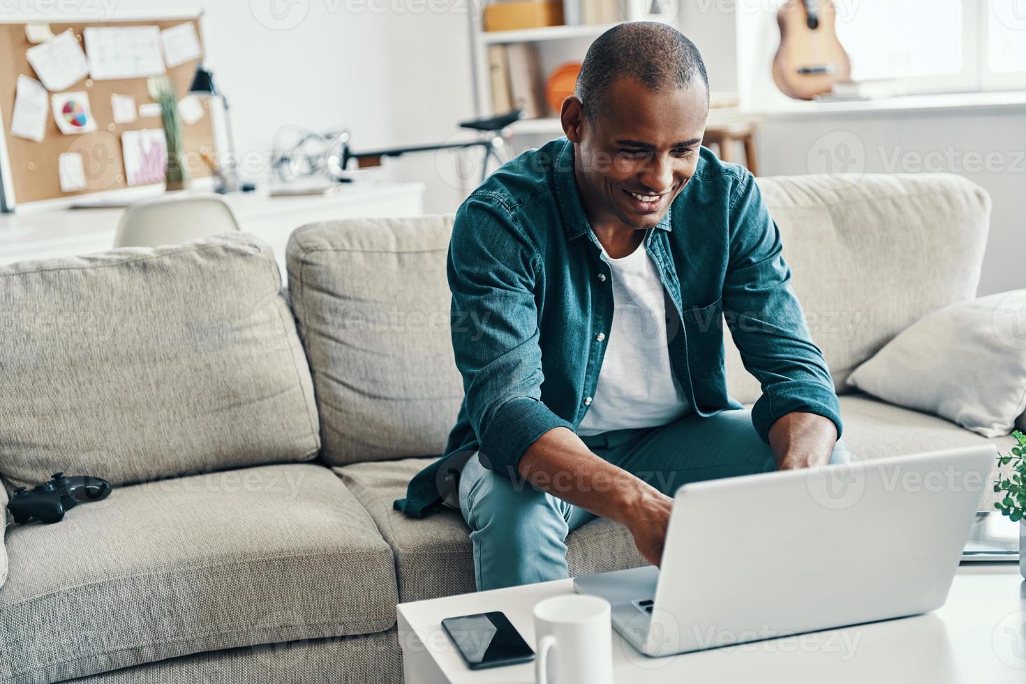 redes sociales. apuesto joven africano usando laptop y sonriendo mientras está sentado adentro foto