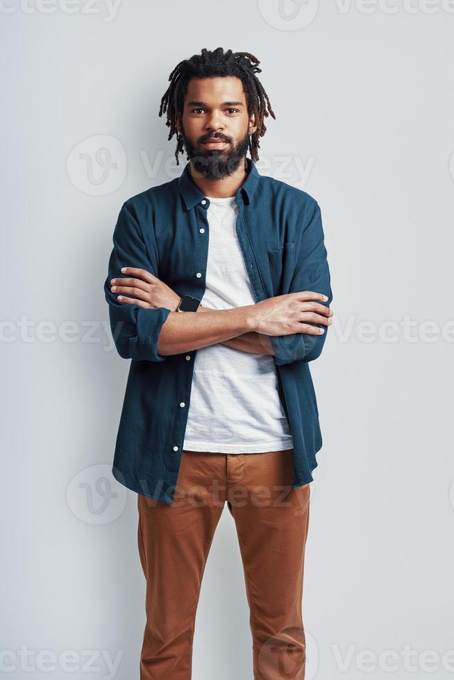 Thoughtful young African man in casual wear looking at camera and keeping arms crossed while standing against grey background photo