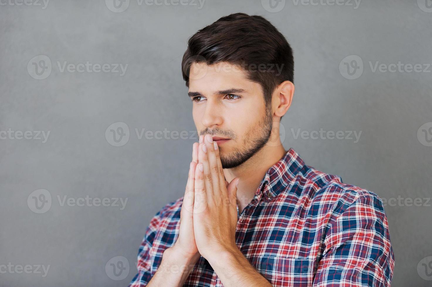 Begging for good. Thoughtful young man holding hands clasped near face and looking away while standing against grey background photo