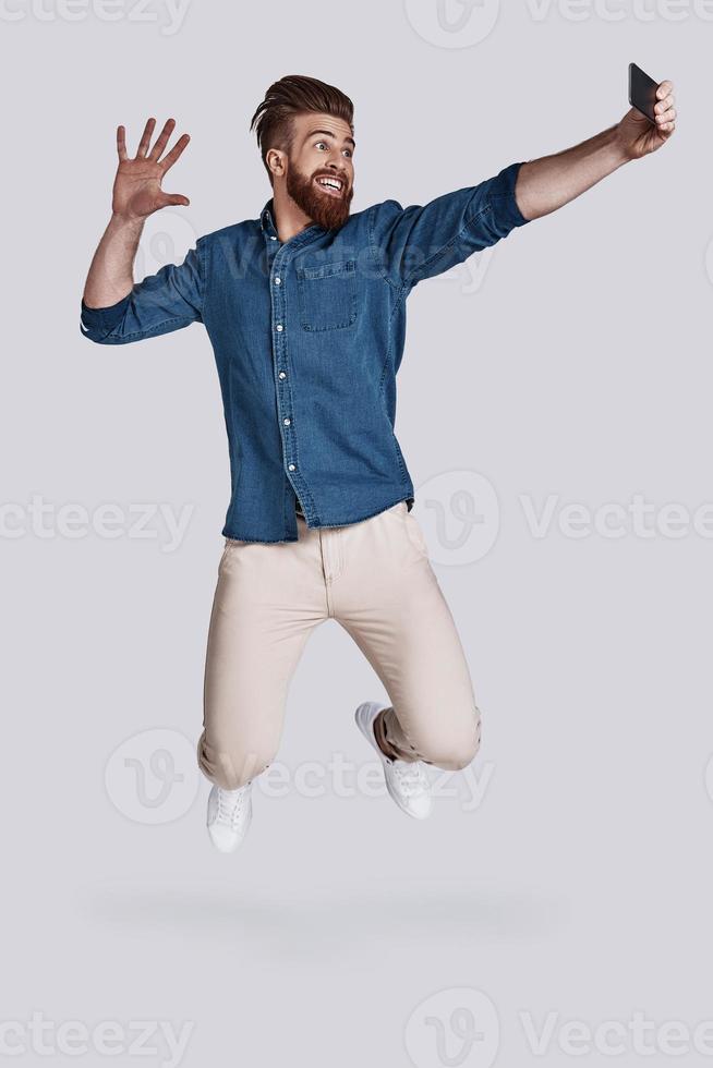 Mid-air selfie.  Full length of handsome young man taking selfie while jumping against grey background photo