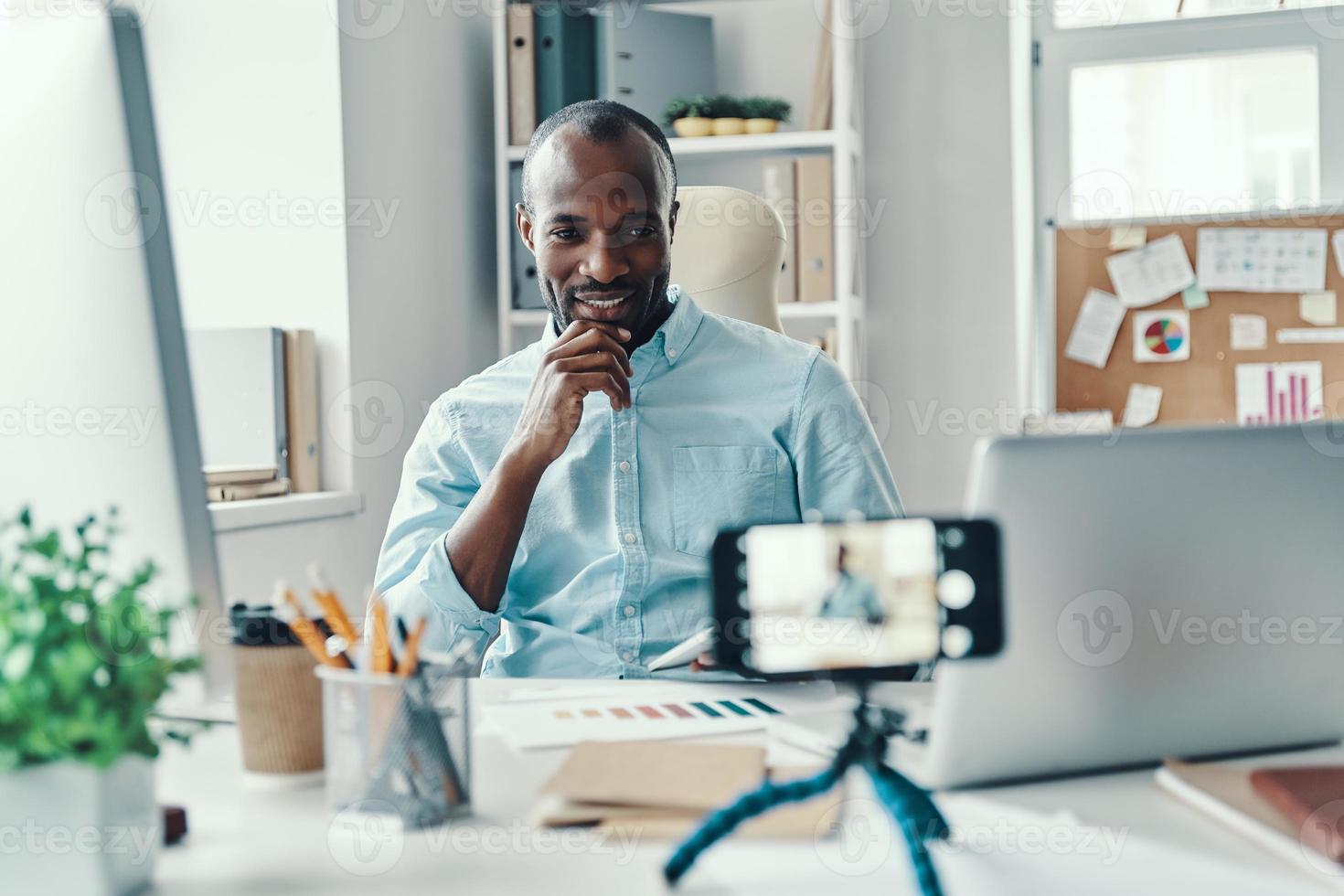 Handsome young African man in shirt telling something and smiling while making social media video photo
