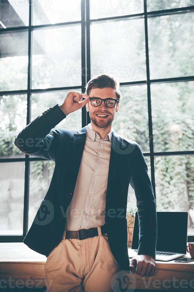 Cheerful young businessman. Confident young man in smart casual wear adjusting his eyeglasses and looking at camera with smile while standing in front of the big window in office or cafe photo