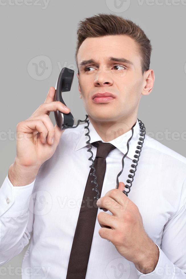 Tired of office work. Depressed young man in shirt and tie talking on the phone and holding telephone wire while standing against grey background photo