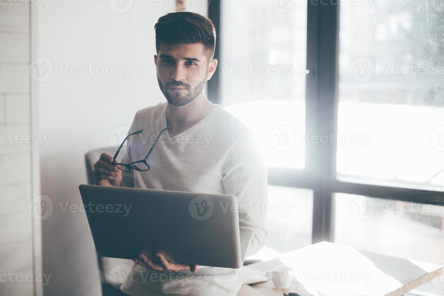 Always ready to help you. Confident young man carrying laptop and eyeglasses while leaning at the desk in office and looking at camera photo