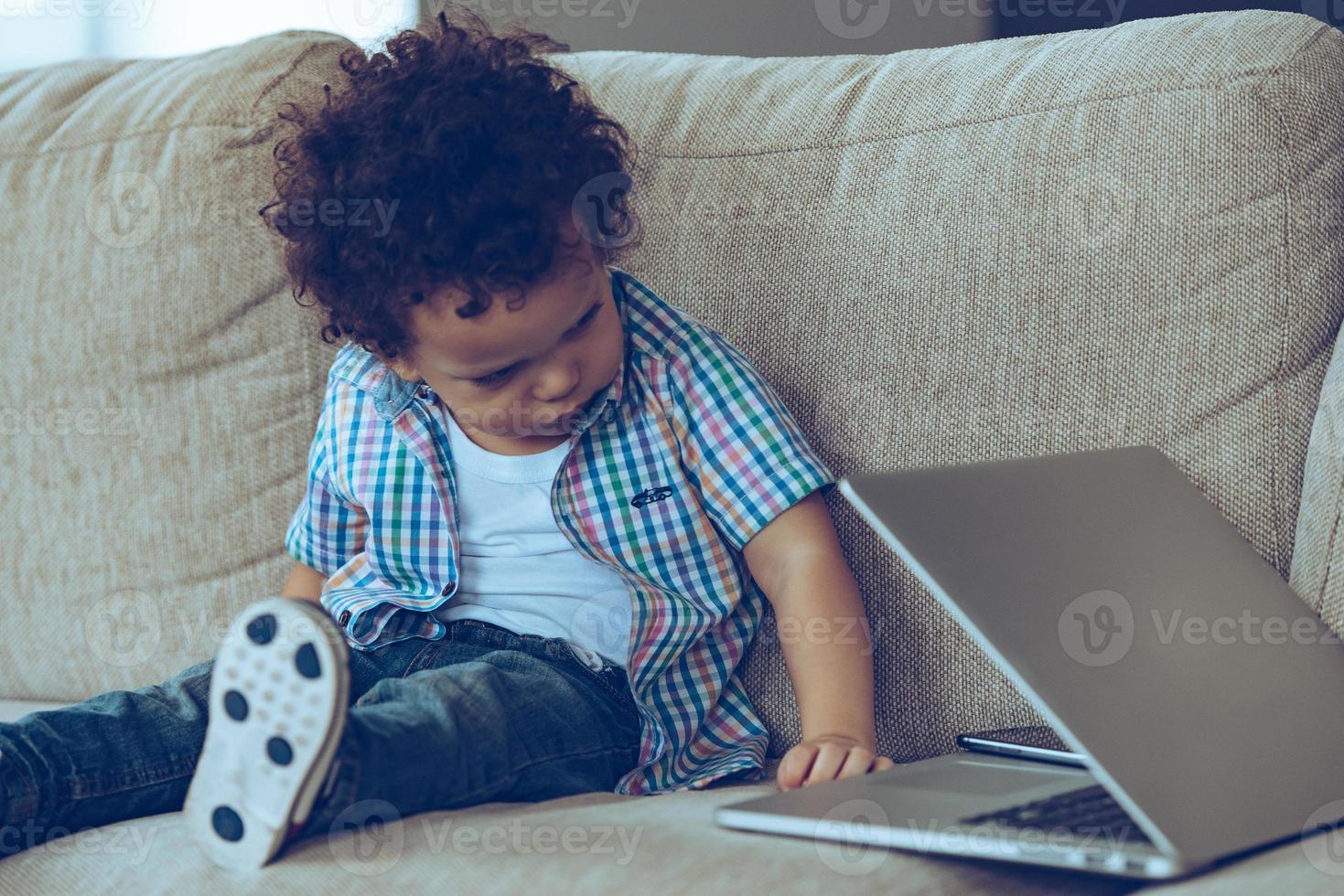 Why is it closed Little African baby boy peeking at his laptop while sitting on the couch at home photo
