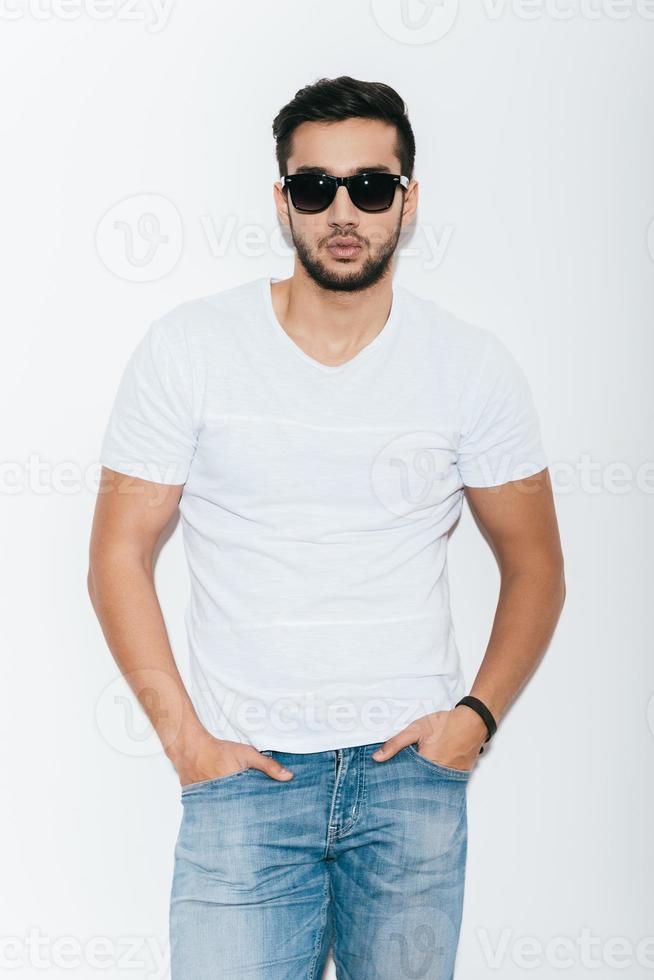 Feeling confident in my skin. Handsome young Indian man in sunglasses holding hands in pockets and looking at camera while standing against white background photo