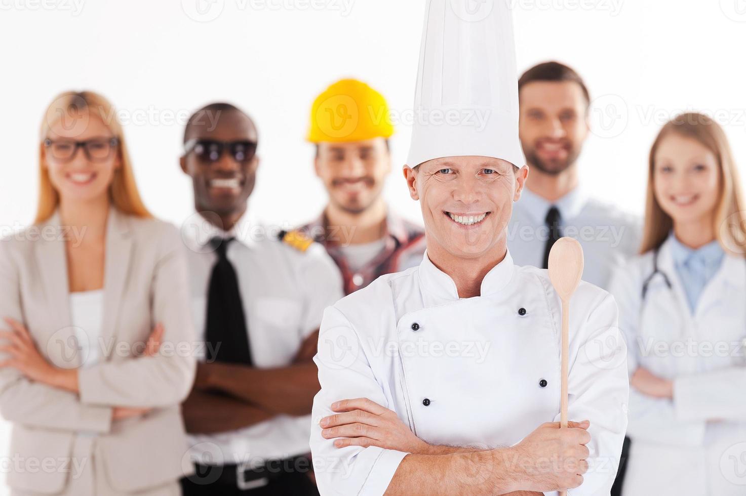 cuando sea grande sere chef chef masculino confiado en uniforme con los brazos cruzados y sonriendo mientras un grupo de personas en diferentes profesiones se paran en el fondo foto