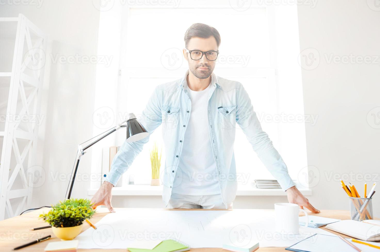 dueño de su lugar de trabajo. apuesto joven con camisa y anteojos mirando a la cámara mientras se inclina en su lugar de trabajo foto