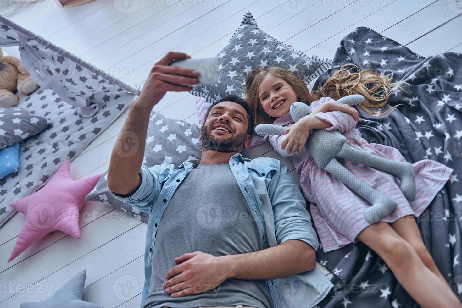 capturar momentos brillantes juntos. vista superior del padre alegre y su pequeña hija tomando selfie mientras están acostados en el suelo juntos en casa foto