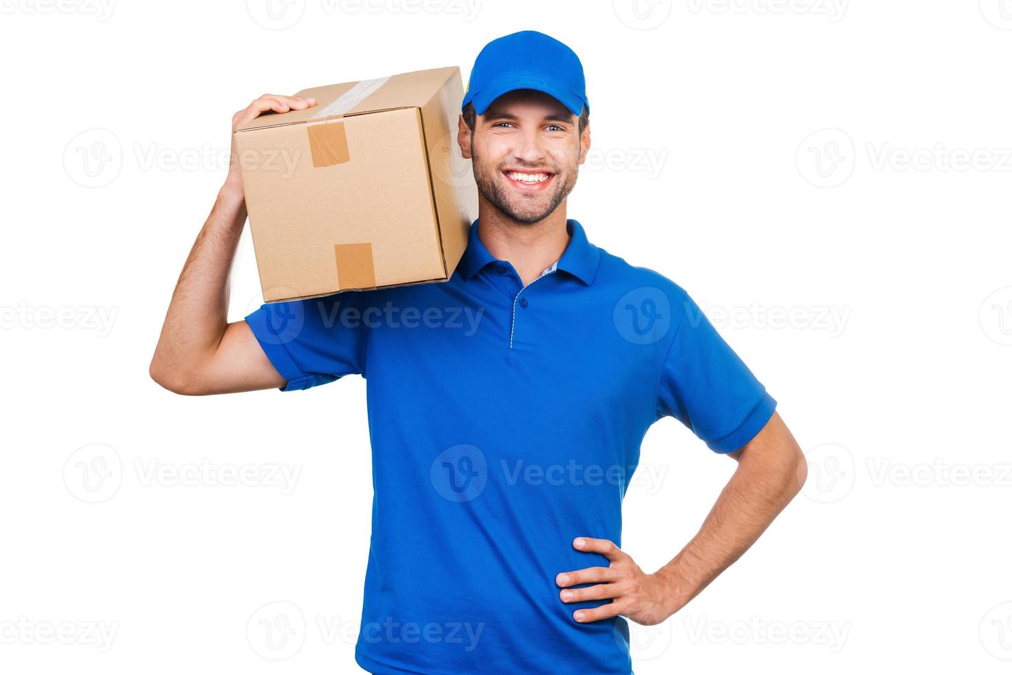 repartidor confiado. mensajero joven alegre que lleva la caja de cartón en el hombro y sonriendo mientras está de pie contra el fondo blanco foto