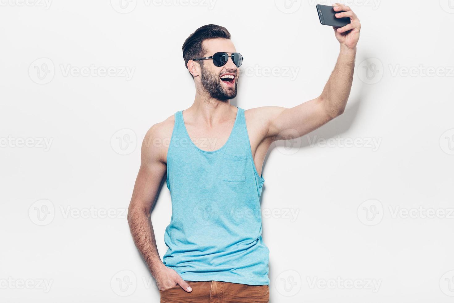 Capturing fun. Happy young handsome man in sunglasses making selfie while standing against white background photo