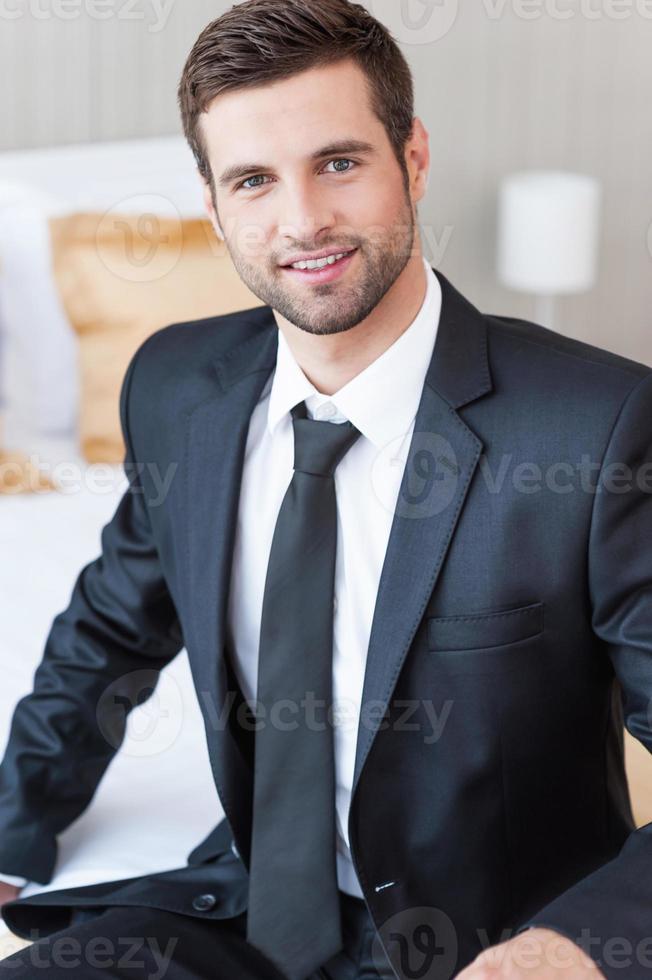 Businessman in hotel room. Confident young businessman in formalwear looking at camera and smiling while sitting on the bed in hotel room photo