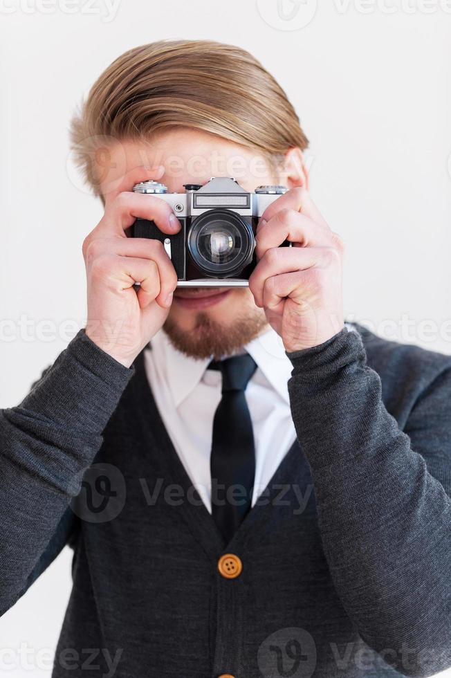 Focusing at you. Young beard man focusing at you with his retro camera while standing against grey background photo