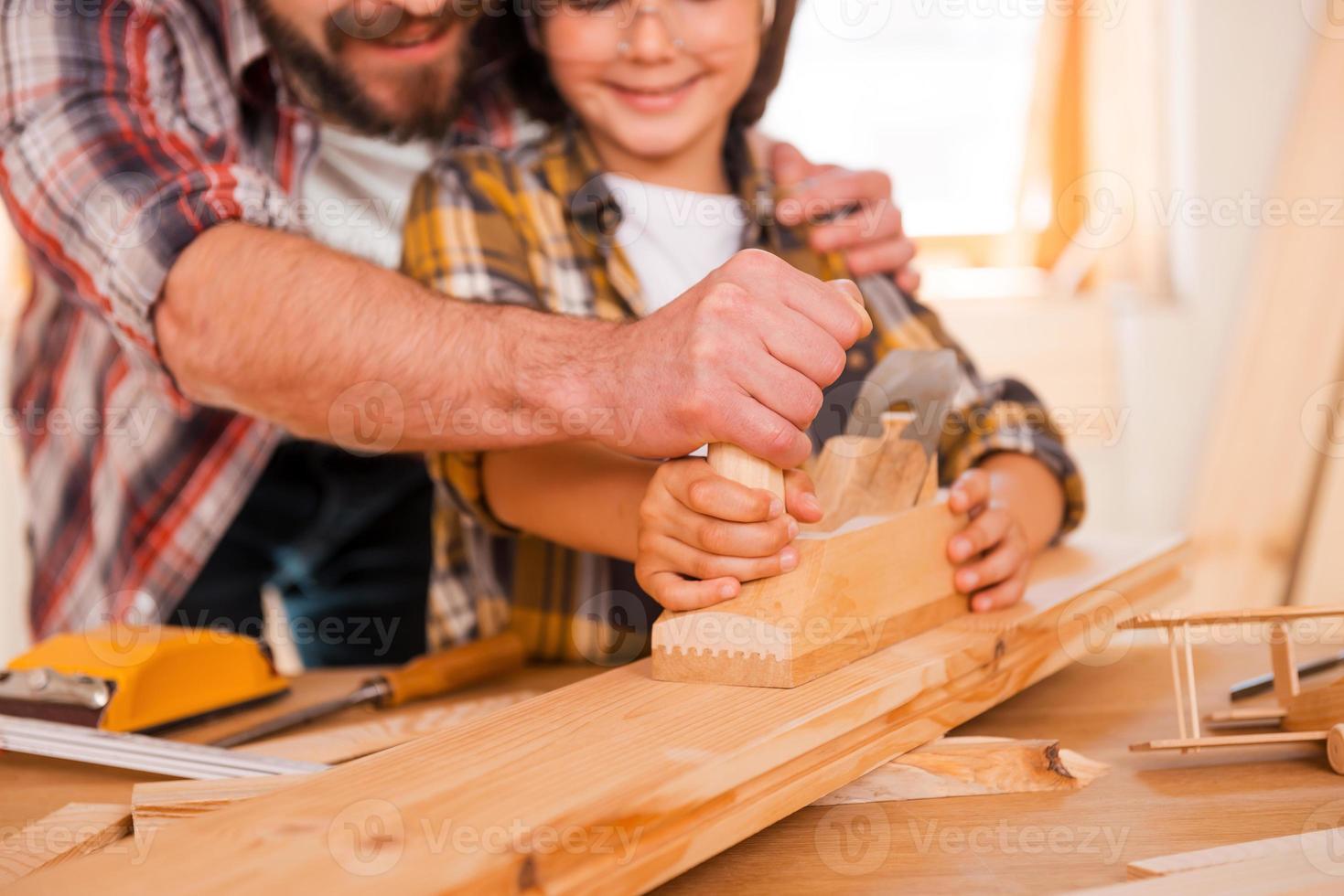 Sharing skills with his son. Close-up of smiling young male carpenter teaching his son to work with wood in his workshop photo