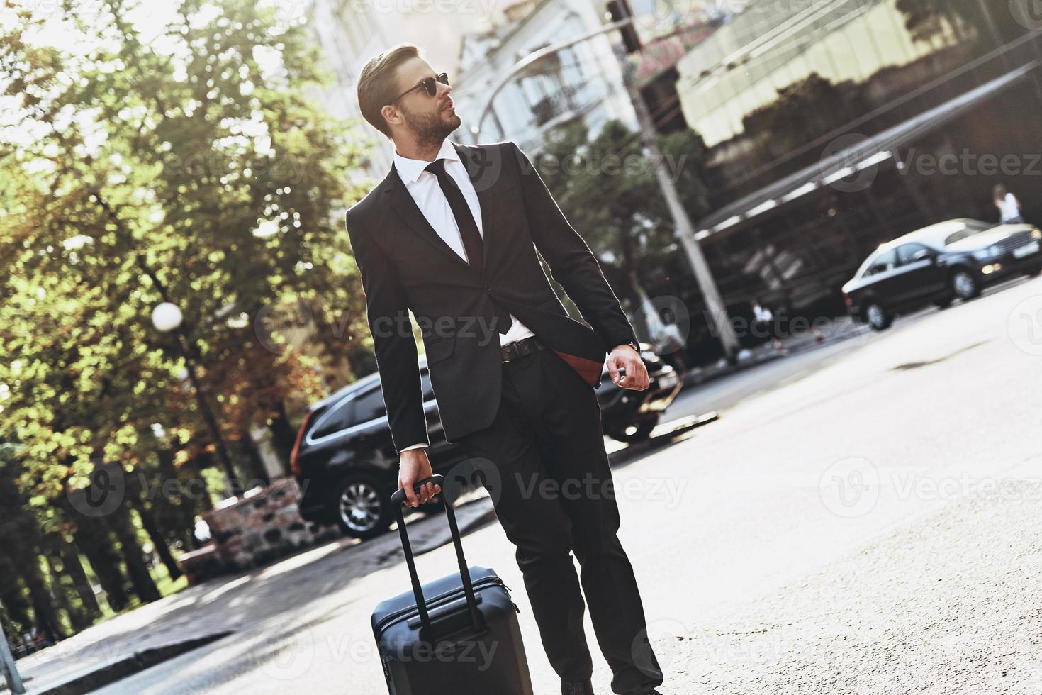 Pure confidence. Handsome young man in full suit pulling luggage while walking outdoors photo