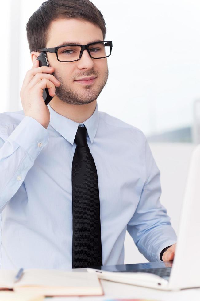 hombre de negocios confiado en el trabajo. un joven confiado con camisa y corbata hablando por teléfono móvil y mirando la cámara mientras se sienta en su lugar de trabajo foto