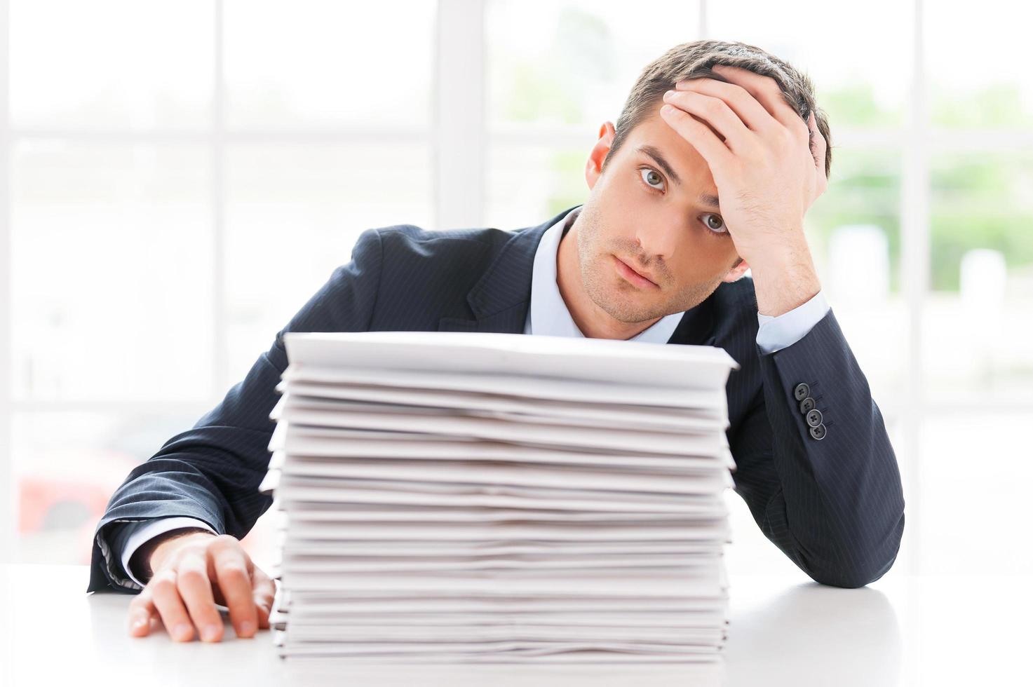 Missing deadlines. Depressed young man in shirt and tie looking at camera and holding head in hand while sitting at the table with stack of documents laying on it photo