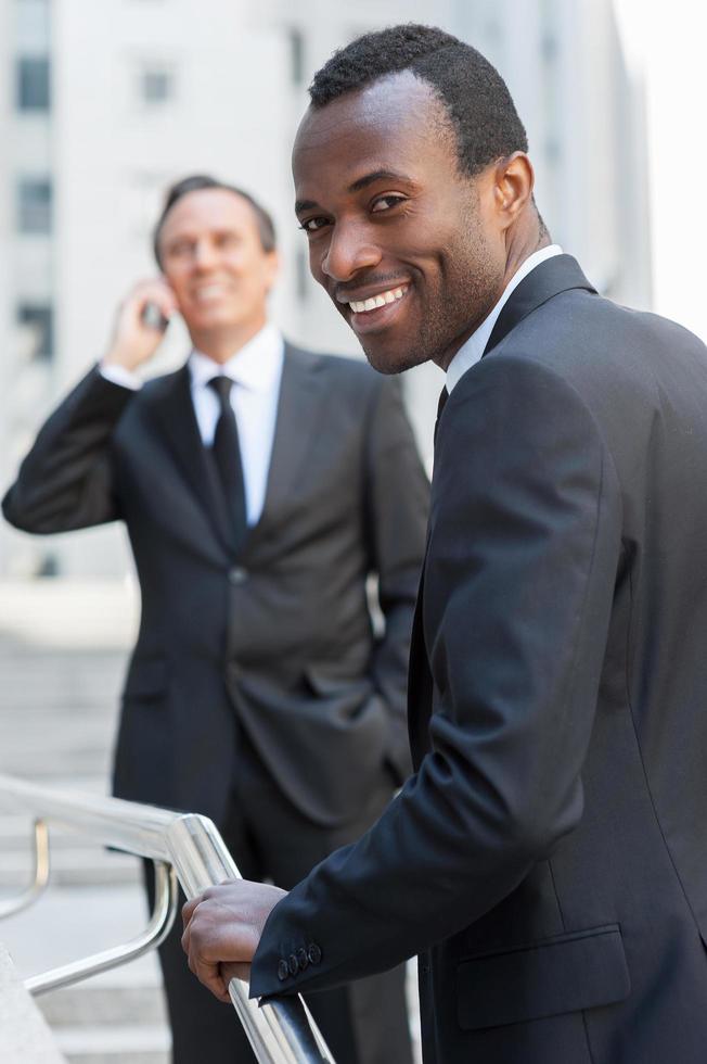 Confident businessman. Cheerful young African man in formalwear moving up by stairs and looking over shoulder while senior man talking on the mobile phone in background photo