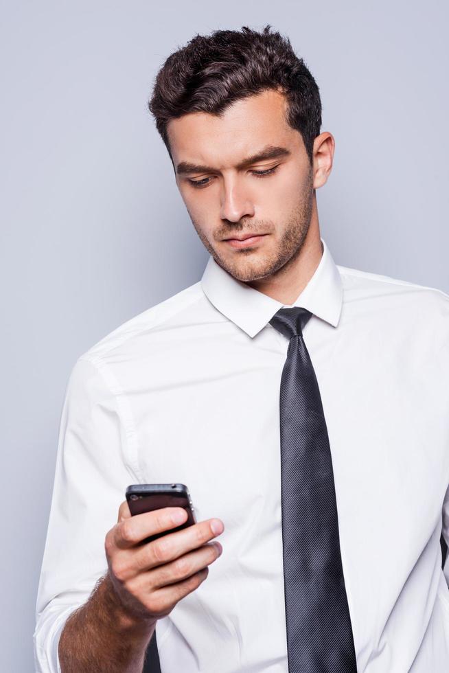 Typing business message. Confident young man in shirt and tie holding mobile phone and looking at it while standing against grey background photo