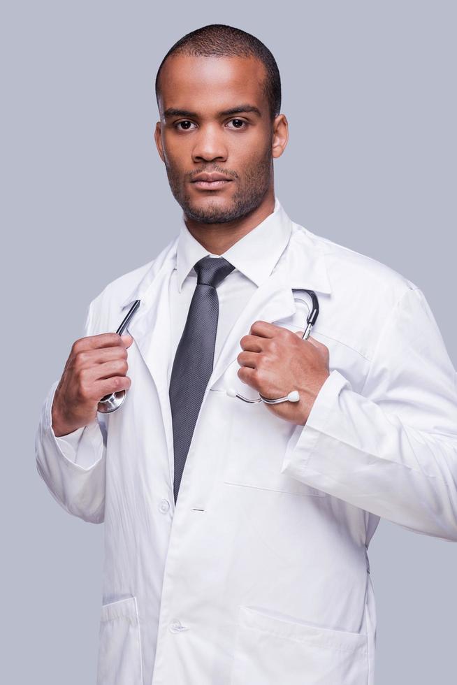 Confident medical expert. Confident African doctor looking at camera and adjusting his stethoscope while standing against grey background photo