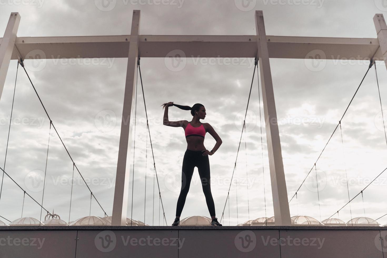 Full of energy. Full length of modern young woman in sports clothing keeping hand on hip and touching her hair while standing outdoors photo