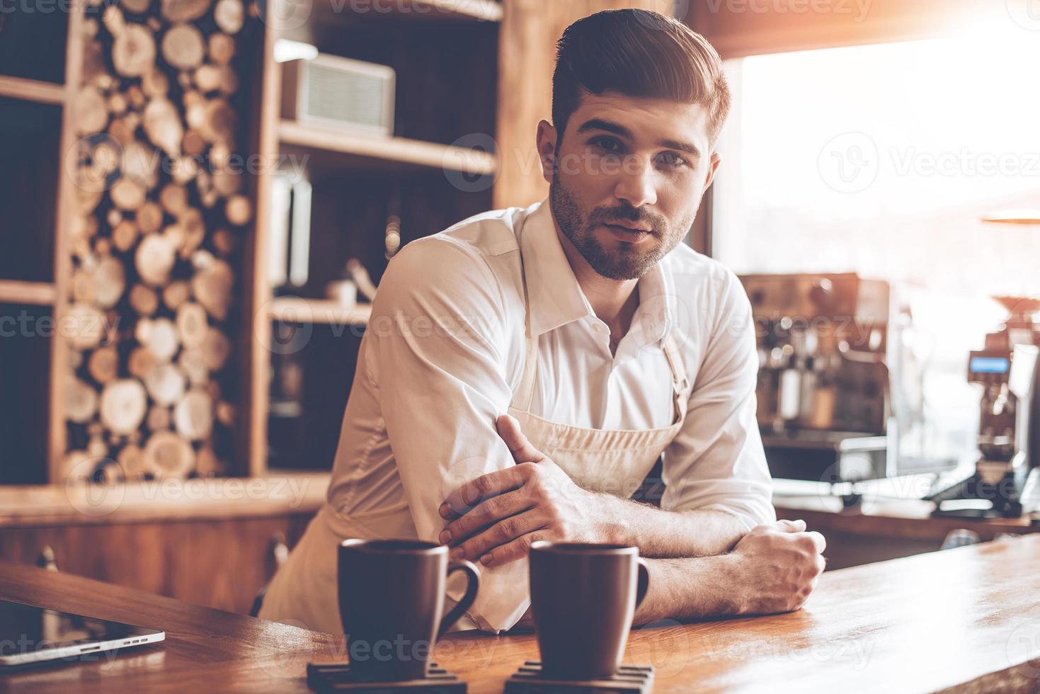 Confident barista. Young handsome man in apron looking at camera while leaning to bar counter at cafe photo