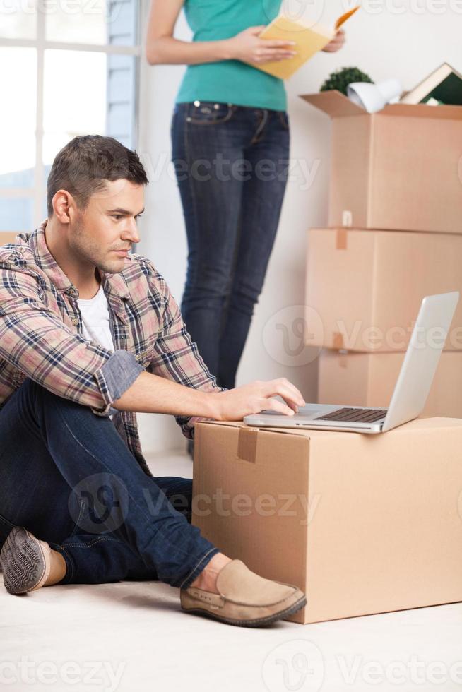 Surfing the net in a brand new house. Confident young man sitting on the floor and working on laptop while woman unpacking a carton box on the background photo