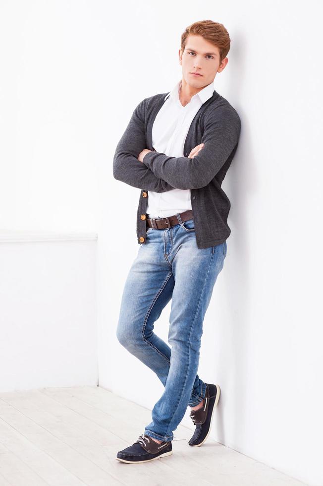 Confident handsome. Full length of handsome young man keeping arms crossed and looking at camera while leaning at the wall photo