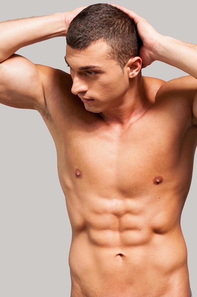 Masculinity. Handsome young shirtless man holding hands behind head and looking away while standing against grey background photo